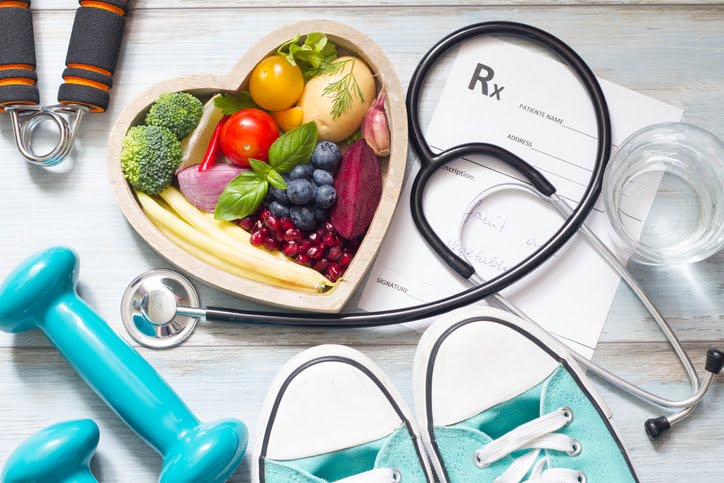 A heart-shaped bowl of produce on a table surrounded by a glass, weights, sneakers, a stethoscope, and a prescription script.