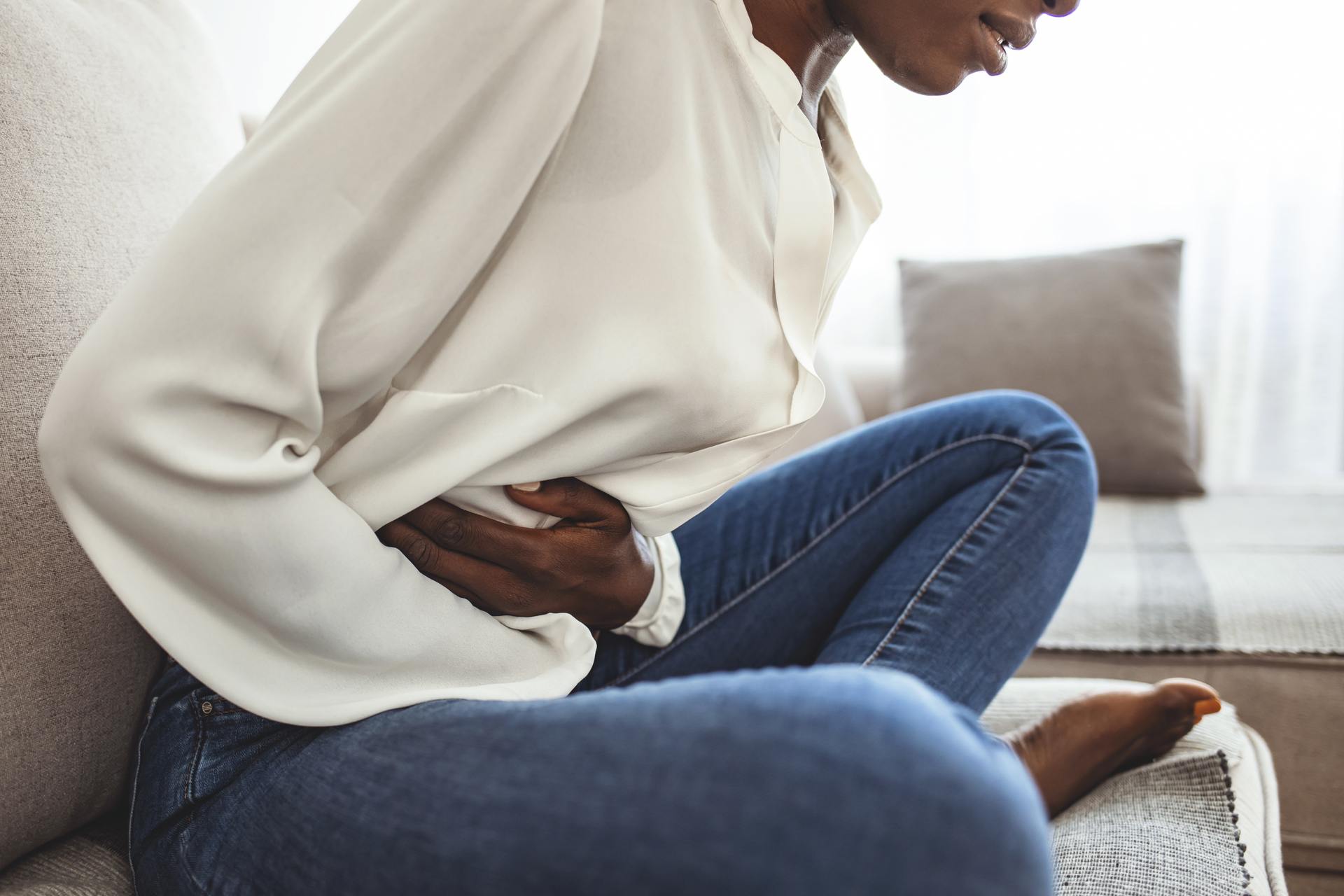 What is Dumping Syndrome A woman holding her stomach in pain wearing an off-white blouse and blue jeans sitting on the couch.