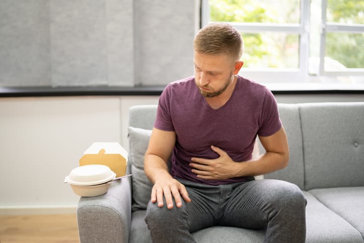 A man in a purple shirt holding his stomach experiencing acid reflux after eating spicy takeout food on his grey couch. 