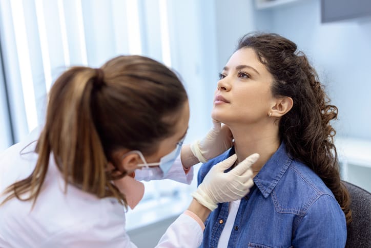 A female doctor wearing a mask examines the throat of a young woman for thyroid disease at a clinic.