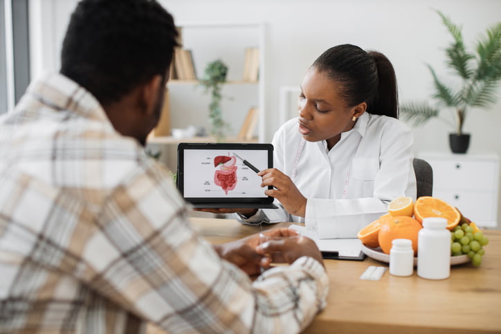 A female doctor in a white coat is holding up a tablet with an illustration of the digestive system to show a male patient in a tan plaid shirt.