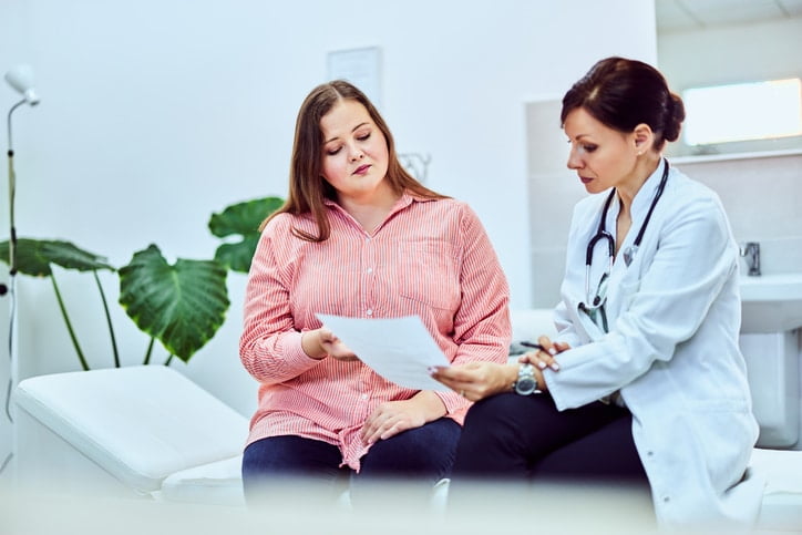 A woman with brown hair in a pink shirt sitting next to a female doctor with dark hair in a white coat holding a piece of paper with a green plant behind them.