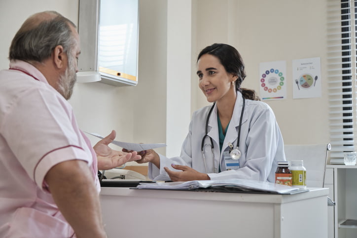 A young female weight loss doctor in a white coat with dark hair sitting at a desk hands a paper to an older male patient in a pink shirt. 